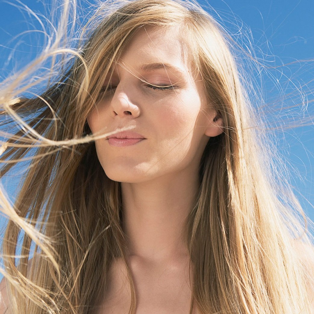 A person with long, light hair closes their eyes as their hair blows gently in the breeze.